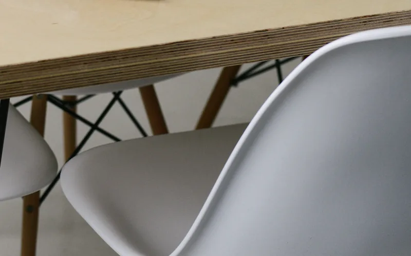 Kante einer Schichtholzplatte, darunter weiße Schalenstühle auf Holz- und Drahtgestellen